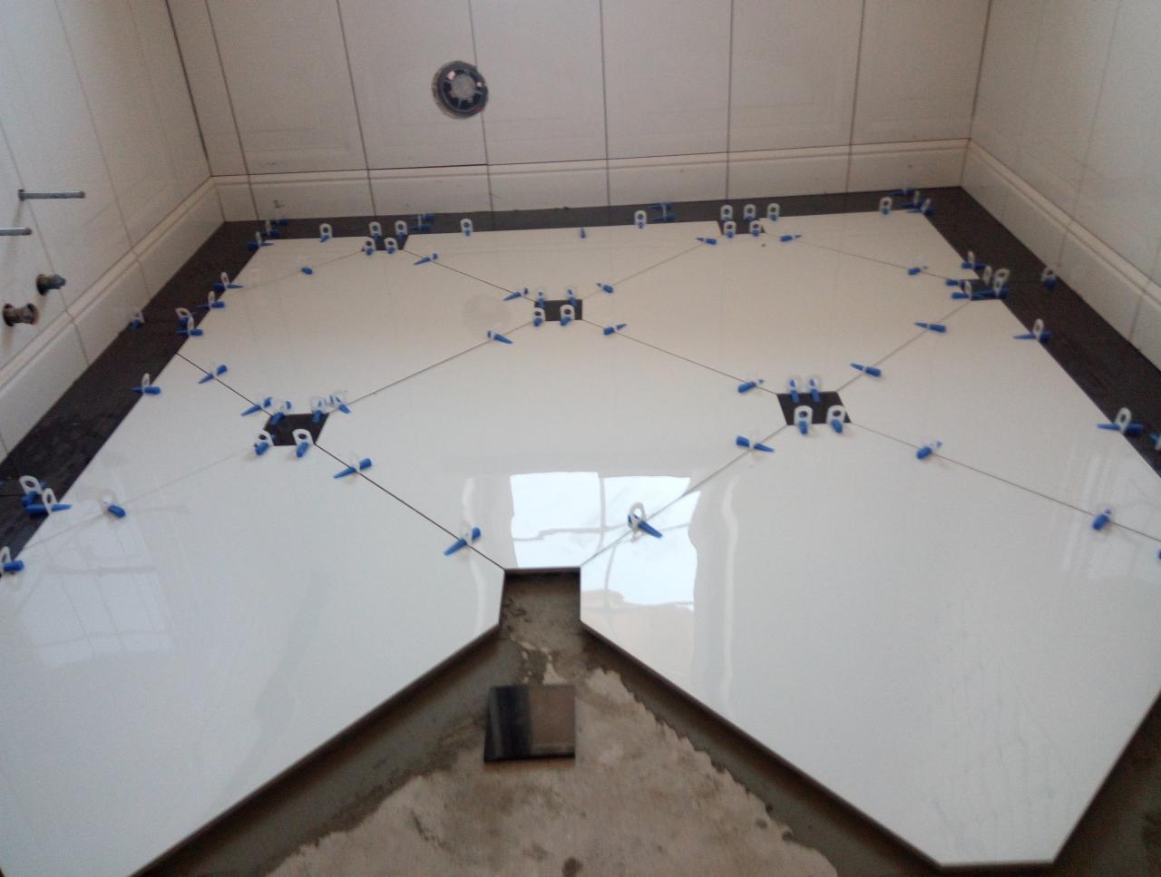 Białe, błyszczące płytki podłogowe ułożone w romby, z czarnymi akcentami na obrzeżach, zabezpieczone niebieskimi klipsami poziomującymi podczas układania w łazience.