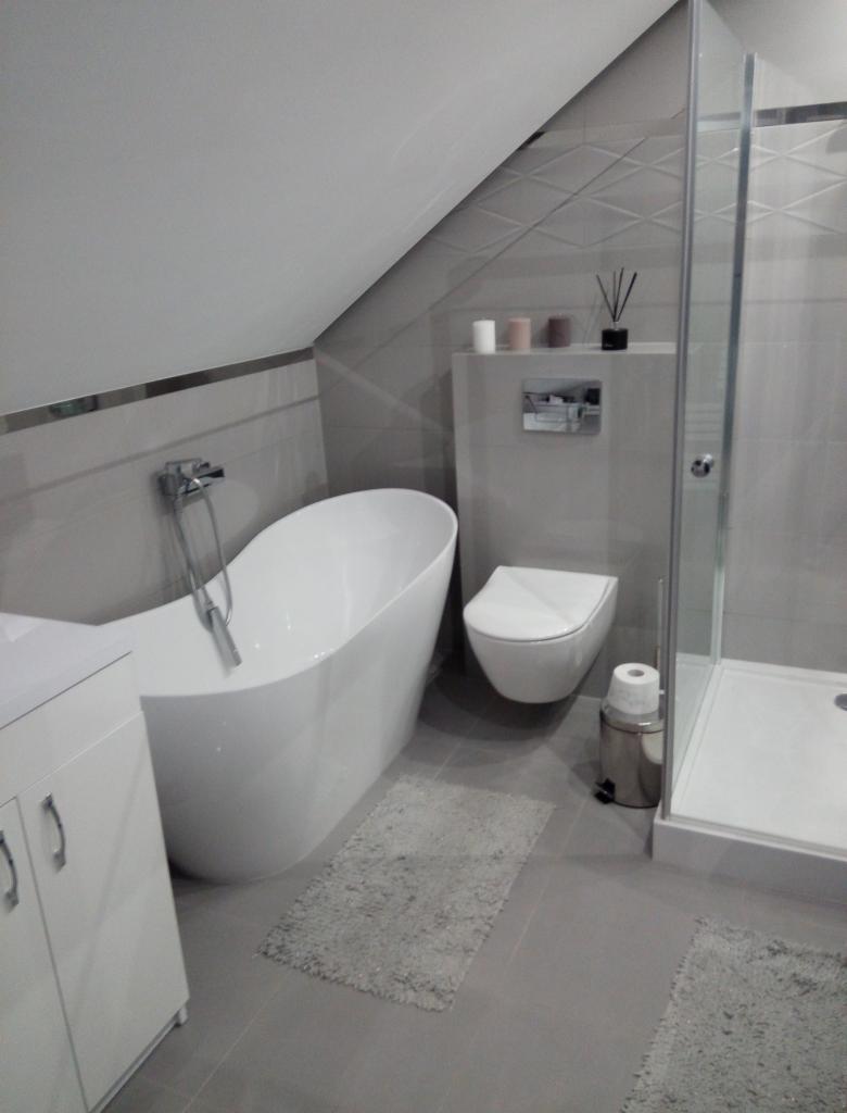 Nowoczesna łazienka na poddaszu z wolnostojącą białą wanną, kabiną prysznicową ze szklanymi drzwiami, podwieszaną toaletą i szarą płytką na ścianach i podłodze.