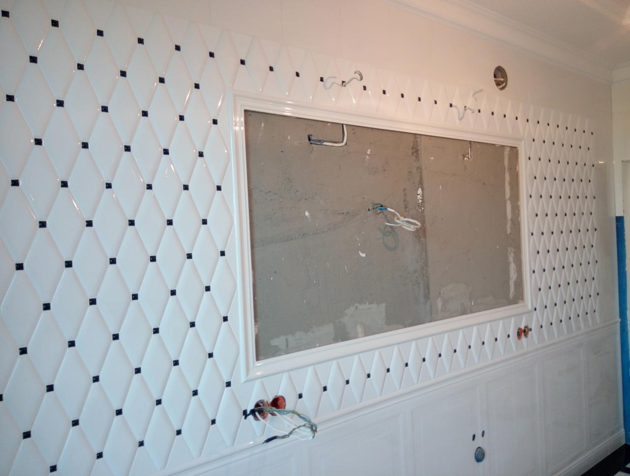 Ściana z białych, błyszczących płytek ułożonych w romby z czarnymi akcentami, obramowanie okna przygotowane do dalszych prac, widoczne przewody elektryczne.