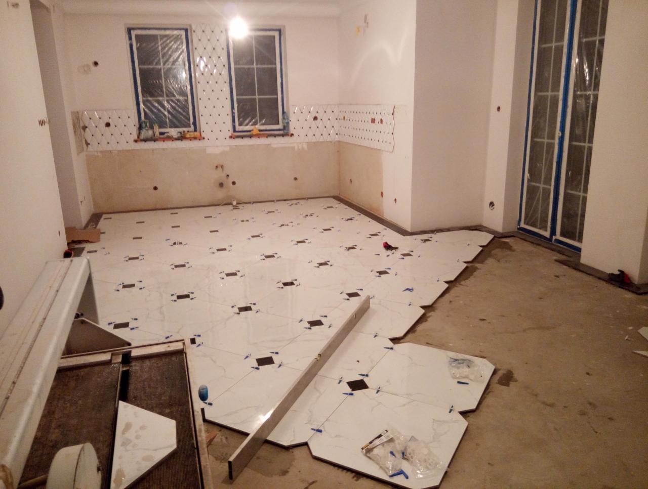 Układanie białych płytek podłogowych z czarnymi wstawkami w pomieszczeniu z przygotowaną ścianą pod zabudowę kuchenną i oknami zabezpieczonymi folią.