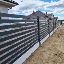 Nowoczesne, poziome przęsła z metalu w kolorze grafitowym tworzą ogrodzenie frontowe wzdłuż nowo wybudowanych domów jednorodzinnych, widoczne samochody i materiały budowlane.