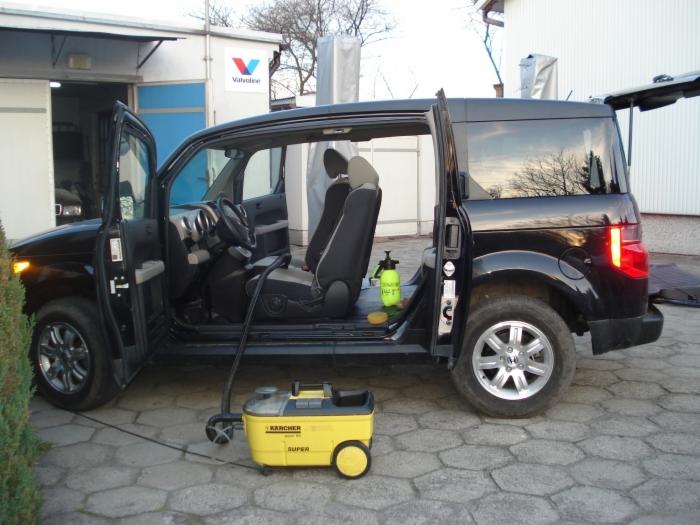 Czarny samochód z otwartymi drzwiami, widoczny odkurzacz parowy Karcher Super i butelka ze spryskiwaczem, sugerujące czyszczenie wnętrza pojazdu.
