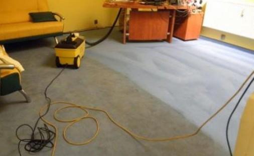 Żółty automat do czyszczenia dywanów na kółkach, widoczny na tle częściowo wyczyszczonej szarej wykładziny w pokoju z żółtymi ścianami i meblami.