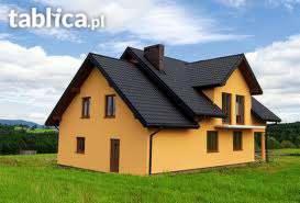 Nowo wybudowany, dwukondygnacyjny dom jednorodzinny z ciemną dachówką, jasną elewacją i brązowymi oknami, na tle zielonego trawnika i błękitnego nieba z chmurami.