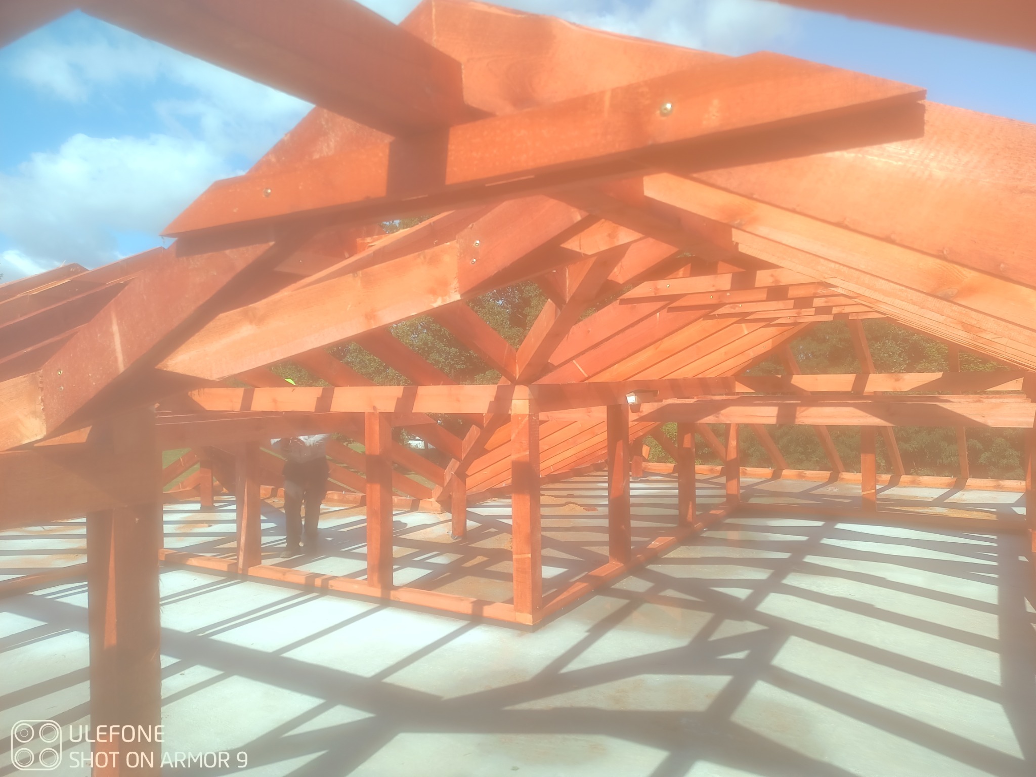 Wnętrze konstrukcji drewnianej dachu w trakcie budowy, widok z dołu na niebo, częściowo widoczna sylwetka pracownika w tle. Elementy złączone śrubami.