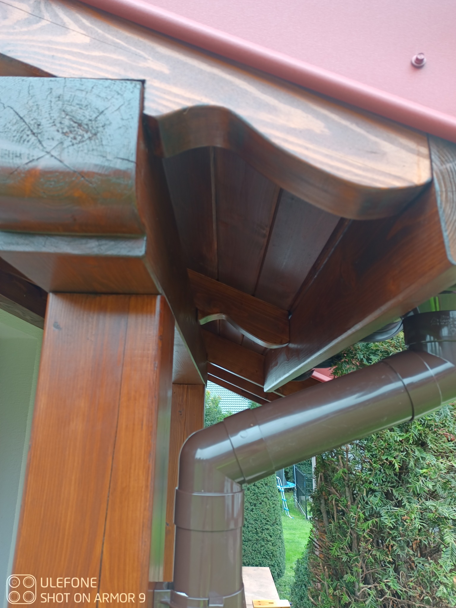 Drewniana konstrukcja z brązową rynną, widok z dołu. Elementy drewniane pomalowane na brązowo, widoczne słoje drewna. W tle zieleń i fragment dachu.