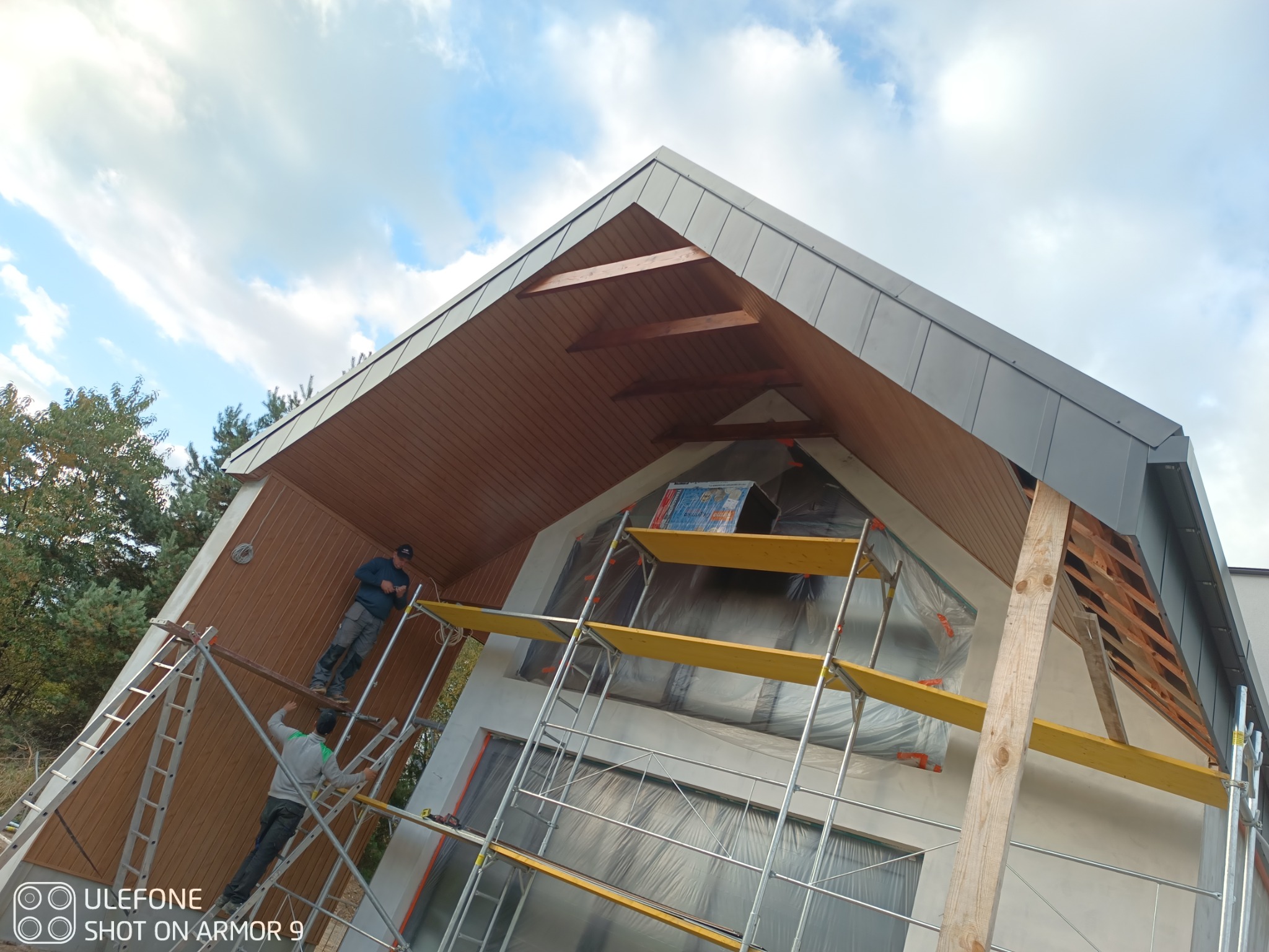 Montaż drewnianej podbitki dachowej na domu jednorodzinnym. Dwóch pracowników na rusztowaniu. Widoczne elementy konstrukcji dachu i fragmenty elewacji zabezpieczone folią.
