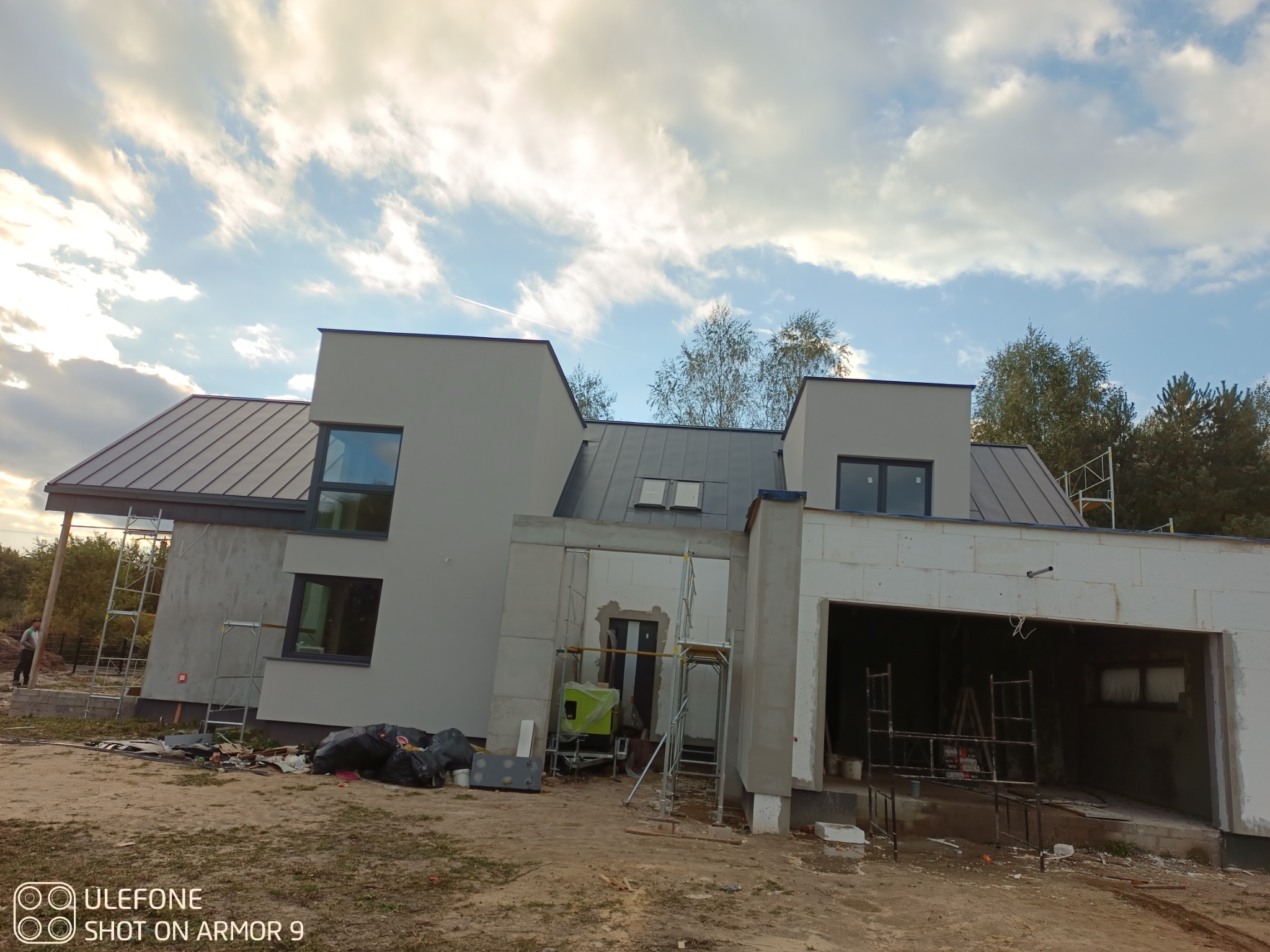 Szary dom jednorodzinny w trakcie budowy, widoczne rusztowania i materiały budowlane. Dach pokryty blachą, okna w ciemnych ramach. Częściowo otynkowana elewacja.