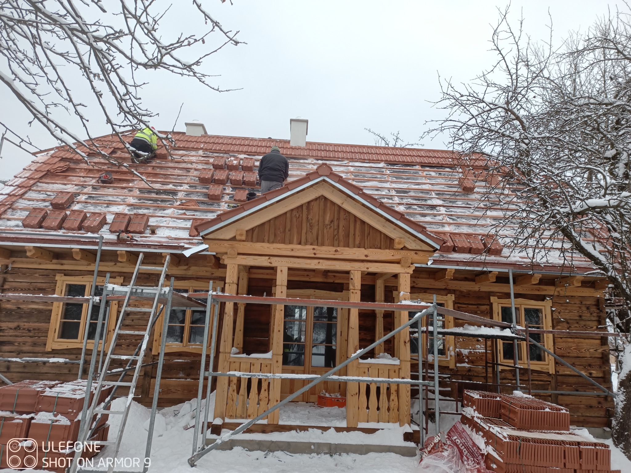 Montaż czerwonej dachówki na drewnianym domu w zimowej scenerii. Widoczni dekarze na dachu, rusztowanie i palety z dachówkami. Praca w toku, zimowy krajobraz.