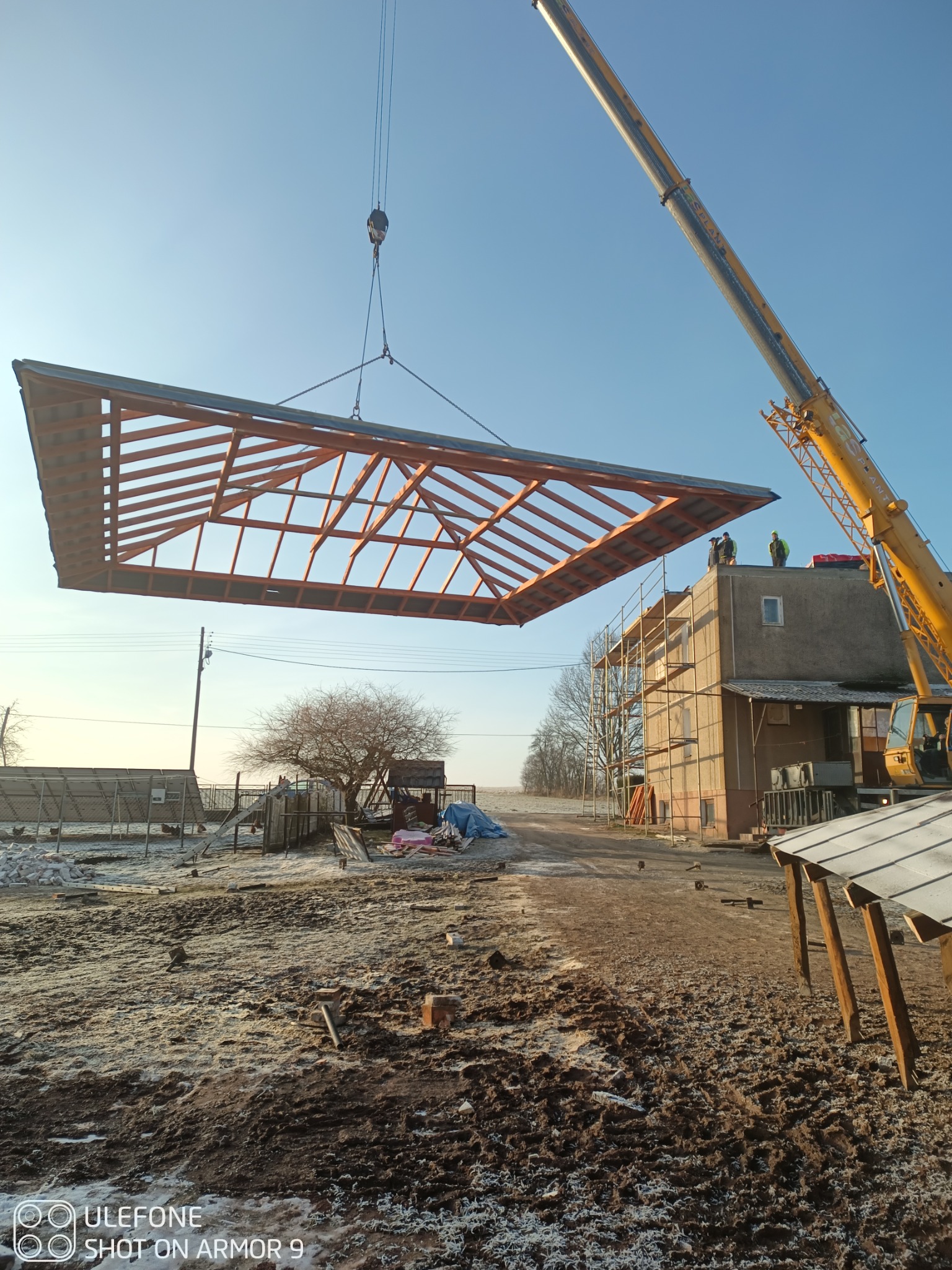 Montaż więźby dachowej za pomocą dźwigu na tle budynku w trakcie budowy. Widoczni pracownicy na dachu. Zimowy krajobraz.