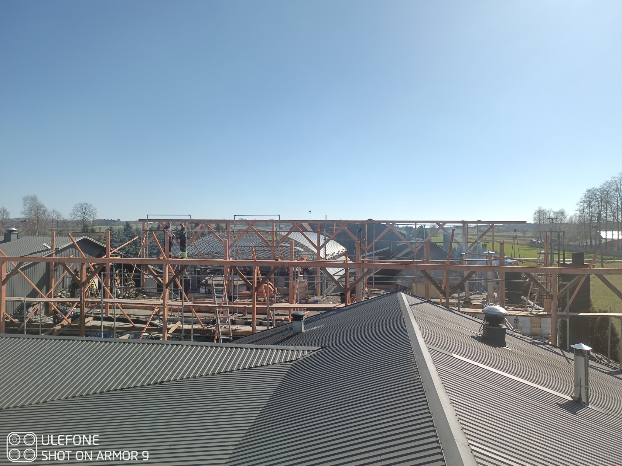 Montaż drewnianej konstrukcji dachu z blachy trapezowej w tle, widok z góry. Ekipa budowlana pracuje na wysokościach przy słonecznej pogodzie.