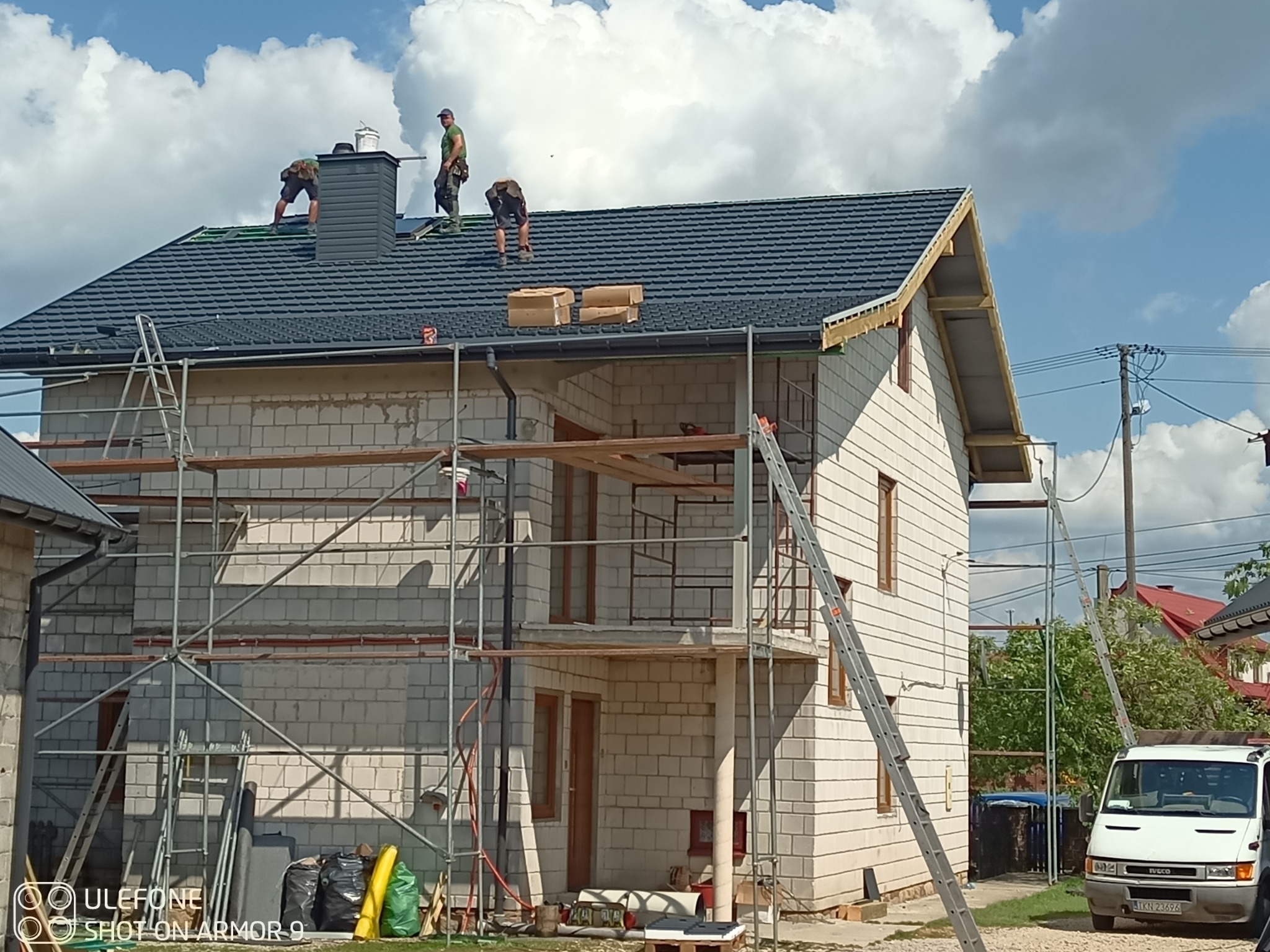 Ekipa dekarzy montuje ciemnoszarą blachodachówkę na nowym domu w trakcie budowy. Widoczne rusztowania i materiały budowlane. Słoneczny dzień, częściowe zachmurzenie.