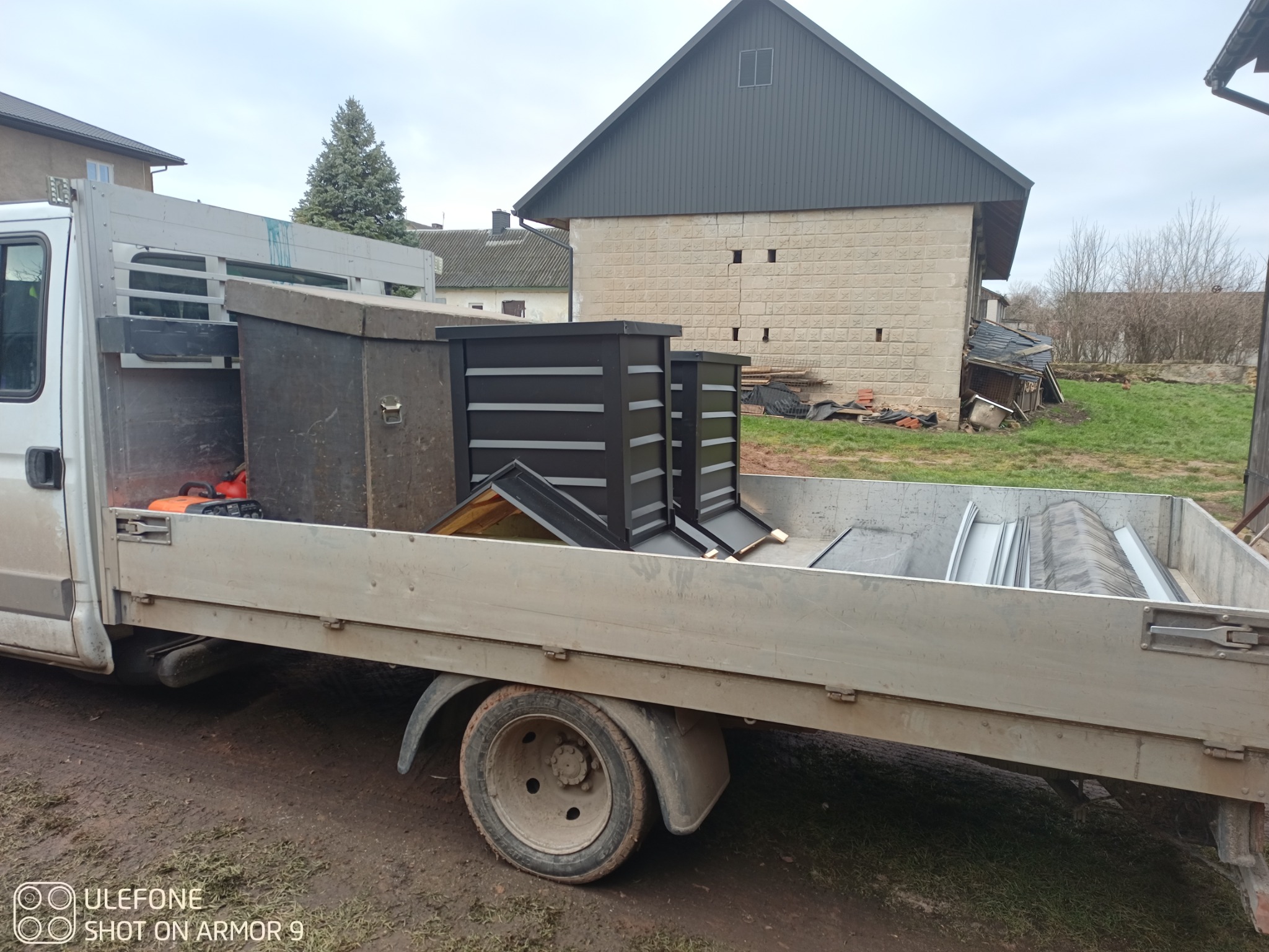 Ciężarówka z blachą dachową i elementami rynien na tle budynku z szarym dachem. Widoczne narzędzia i drabina. Pochmurne niebo.