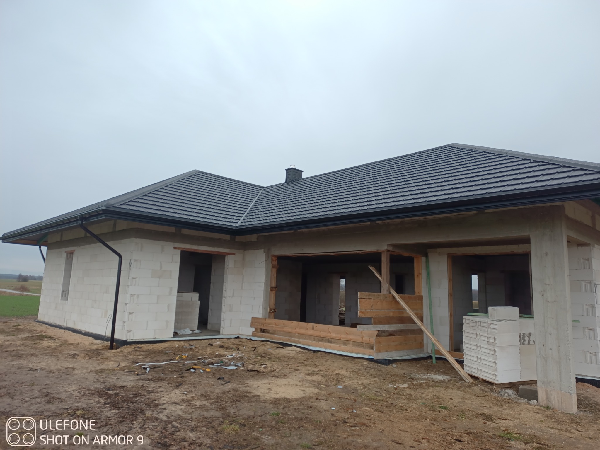 Dom w trakcie budowy z widocznym, nowym dachem z ciemnej dachówki. Elementy konstrukcyjne i materiały budowlane na pierwszym planie. Pochmurne niebo w tle.