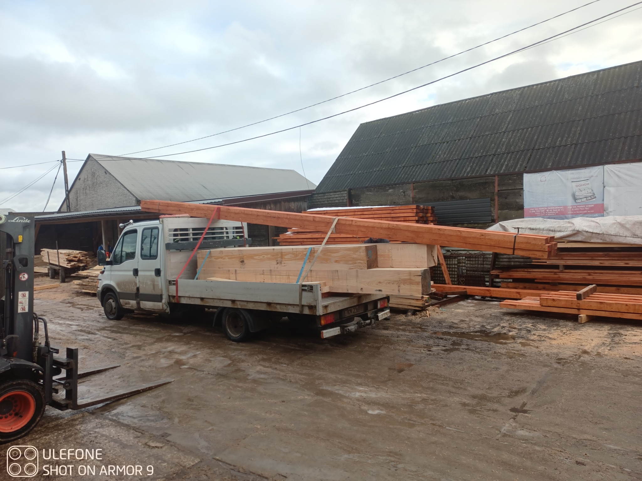 Transport drewna na więźbę dachową w Ruda Maleniecka. Na pace samochodu i obok niego leżą drewniane elementy konstrukcyjne. W tle budynki gospodarcze i wózek widłowy.