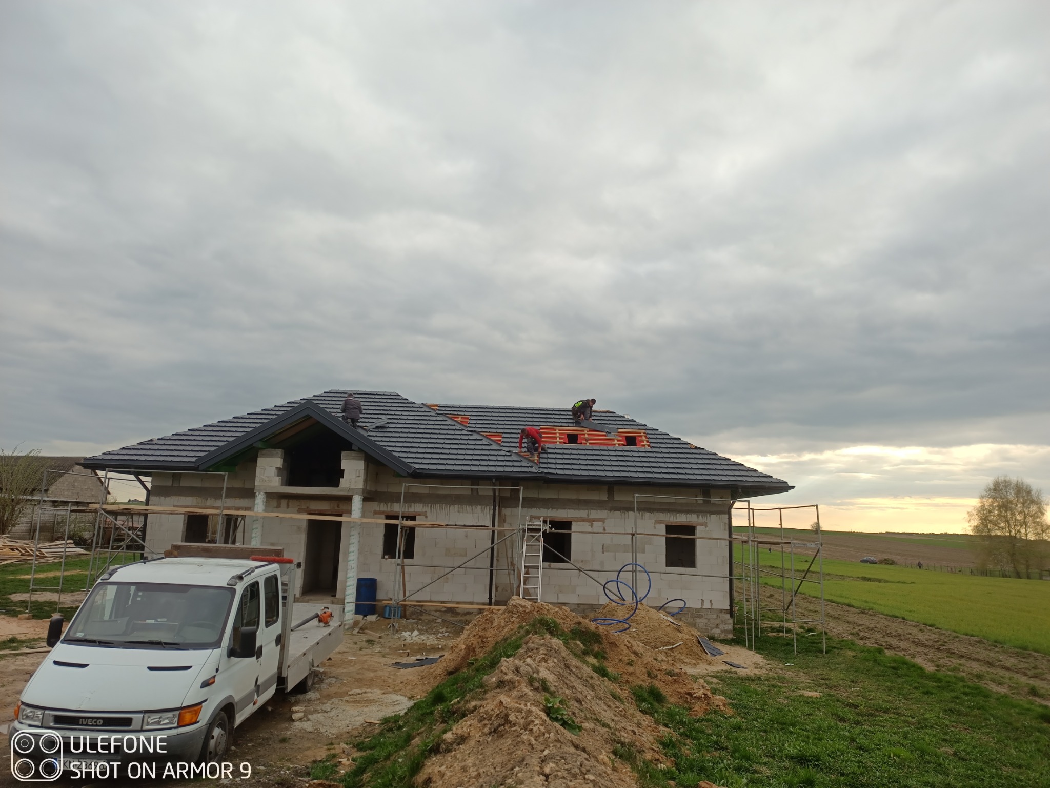 Montaż dachówki na nowym domu w trakcie budowy. Widoczni dekarze na dachu, rusztowanie i samochód dostawczy. Tło stanowią pola i zachmurzone niebo.