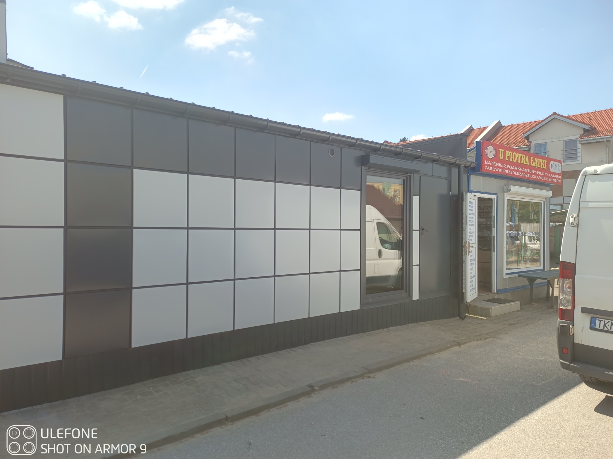 Elewacja budynku wykończona panelami w szachownicę, z widoczną rynną i fragmentem dachu w tle. Obok budka z szyldem 'U Piotra Łatki'.