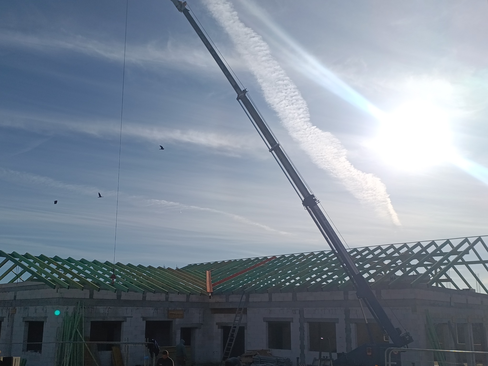 Wznoszenie więźby dachowej z użyciem dźwigu na tle błękitnego nieba z chmurami. Widoczne elementy konstrukcyjne i fragment budynku w stanie surowym otwartym.