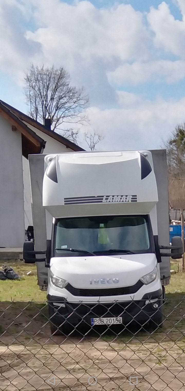 Biały samochód dostawczy marki Iveco z zabudową LAMAR zaparkowany przed domem, widoczny przez siatkę ogrodzeniową.