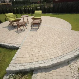 Okrągły taras z jasnej kostki, obrzeże z szarego kamienia, meble ogrodowe z zielonymi poduszkami, trawnik. Ujęcie z góry.