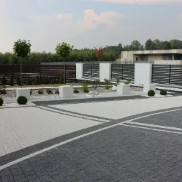 Nowoczesna posesja z geometrycznym wzorem z kostki brukowej w odcieniach szarości i bieli, minimalistyczne ogrodzenie metalowe i dekoracyjne elementy roślinne.