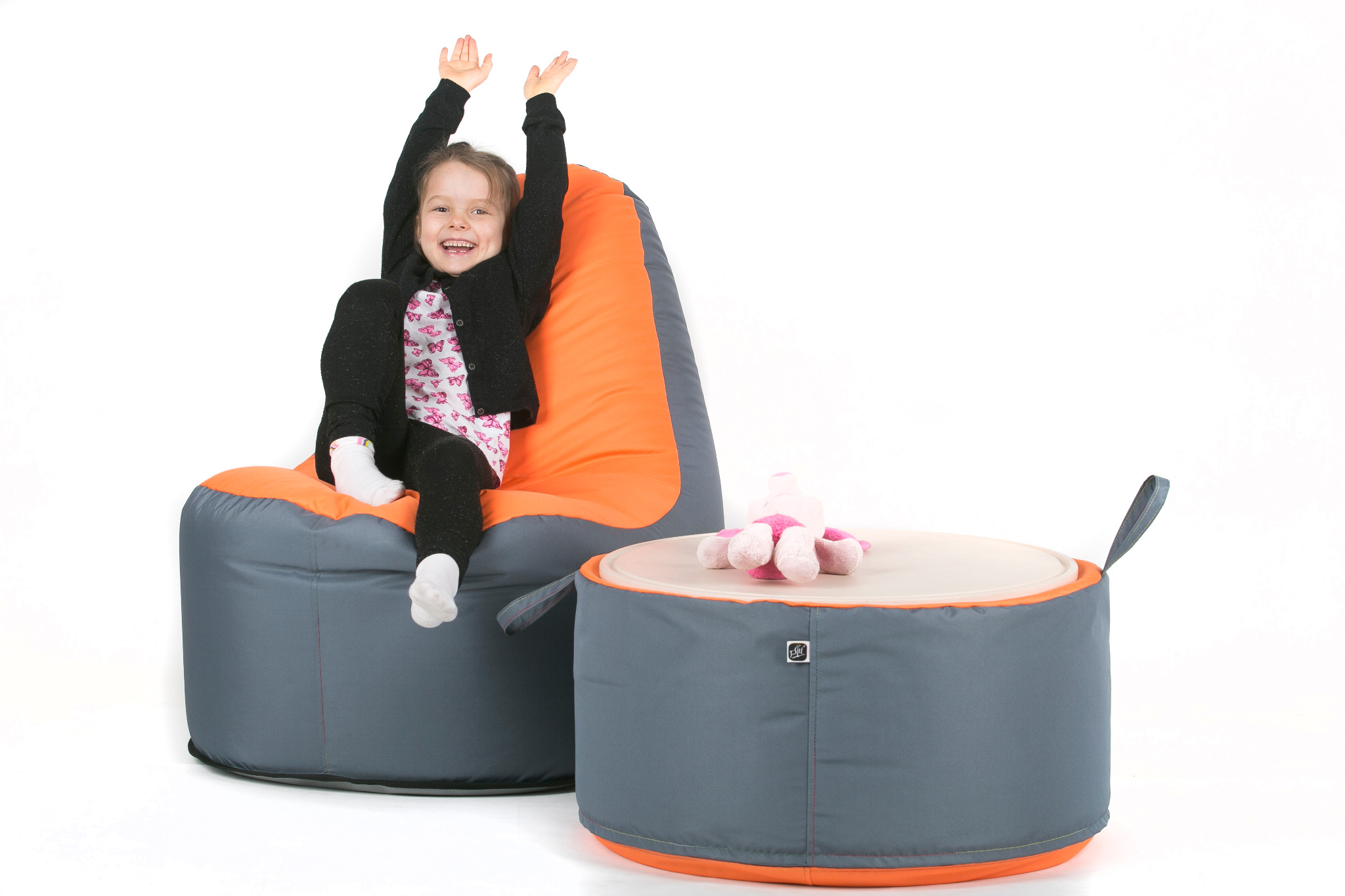 Uśmiechnięta dziewczynka siedzi na szaro-pomarańczowym worku sako, obok stoi pufa ze schowkiem, na której leży pluszowy królik, białe tło.