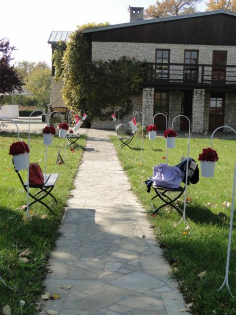 Kamienny chodnik prowadzący do kamiennego domu, ozdobiony białymi stojakami z czerwonymi kwiatami w białych doniczkach, ustawionymi wzdłuż ścieżki, z krzesłami i torebkami na nich.