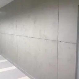 Szara ściana wykończona panelami z płyty kartonowo-gipsowej, z widocznymi łączeniami i listwą przypodłogową, w jasnym korytarzu z kasetonowym sufitem.