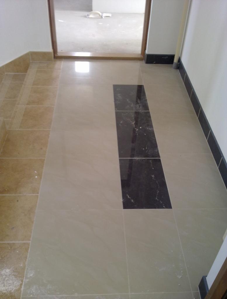 Ułożona podłoga z płytek w odcieniach beżu z wstawką z ciemnych, marmurkowych płytek, widoczne listwy przypodłogowe i próg drzwiowy.