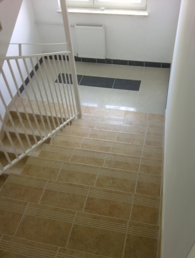 Schody wyłożone płytkami ceramicznymi w odcieniach beżu, z białymi barierkami i grzejnikiem w tle, widoczne czarne akcenty na podłodze przy ścianie.