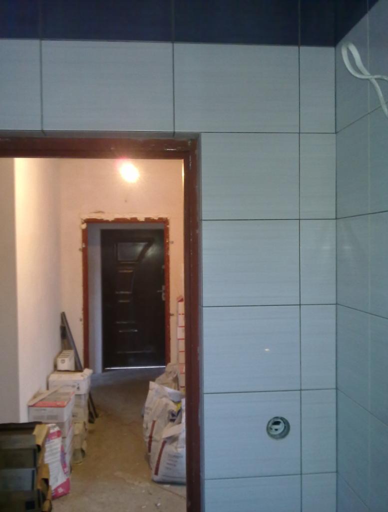 Świeżo ułożone, jasnoszare płytki w łazience, widoczny otwór na gniazdko elektryczne, niewykończone obramowanie drzwi i worki z zaprawą w tle.