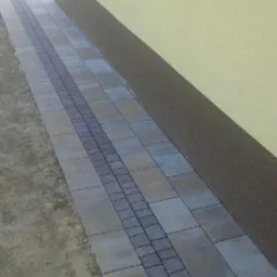 Ułożona kostka brukowa wzdłuż ściany budynku z dwoma odcieniami elewacji. Centralnie pas z mniejszej kostki. Widoczny grunt.