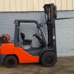 Pomarańczowo-szary wózek widłowy Toyota SAS 35 z butlą gazową z tyłu, stojący na tle blaszanej ściany garażu.