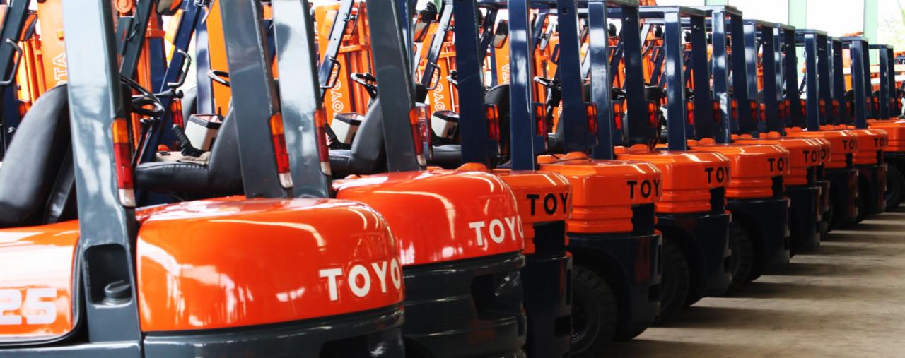 Rząd nowych, pomarańczowo-czarnych wózków widłowych marki Toyota ustawionych w hali magazynowej, widok z niskiej perspektywy na przód pojazdów.
