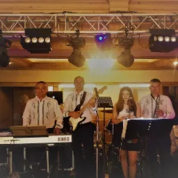 Czteroosobowy zespół muzyczny na scenie podczas występu, mężczyźni w koszulach z haftem, kobieta śpiewa do mikrofonu, widoczne instrumenty: gitara elektryczna, saksofon, keyboard Korg, oświetlenie...