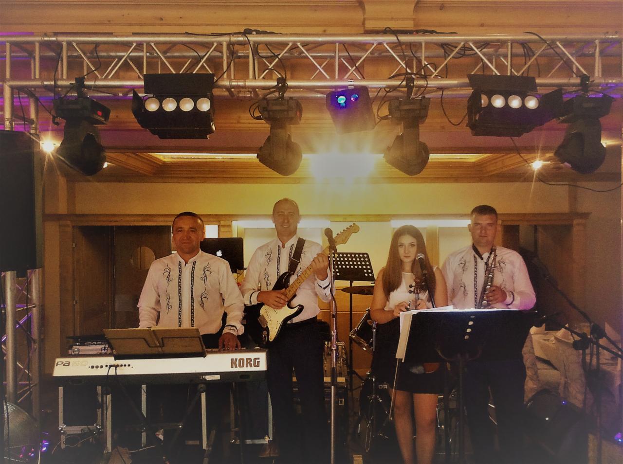 Czteroosobowy zespół muzyczny na scenie podczas występu, mężczyźni w koszulach z haftem, kobieta śpiewa do mikrofonu, widoczne instrumenty: gitara elektryczna, saksofon, keyboard Korg, oświetlenie...