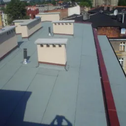 Widok z góry na płaski dach pokryty szarą papą termozgrzewalną z kominami w beżowym kolorze. W tle zabudowa miejska w słoneczny dzień.
