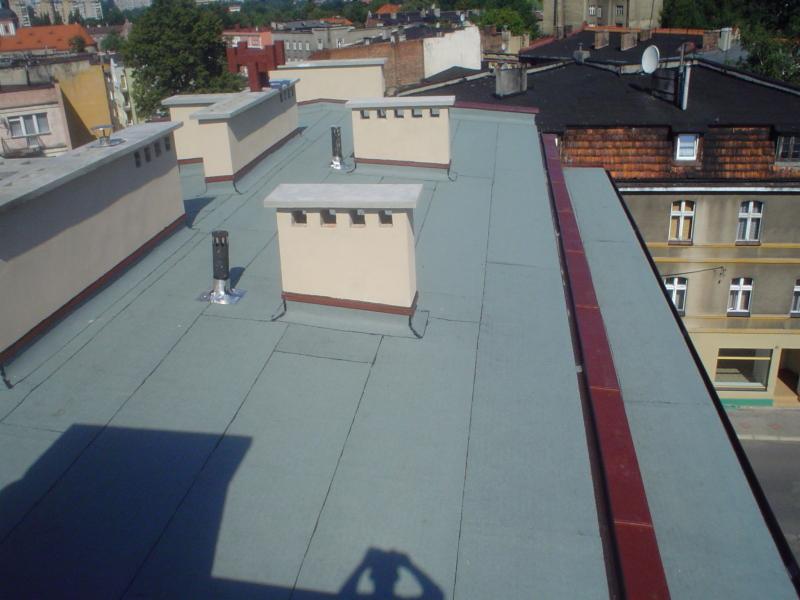 Widok z góry na płaski dach pokryty szarą papą termozgrzewalną z kominami w beżowym kolorze. W tle zabudowa miejska w słoneczny dzień.
