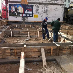 Wylewanie fundamentów z betonomieszarki w Człuchowie. Dwóch pracowników budowlanych w trakcie pracy. Szalunki drewniane i zbrojenie widoczne.