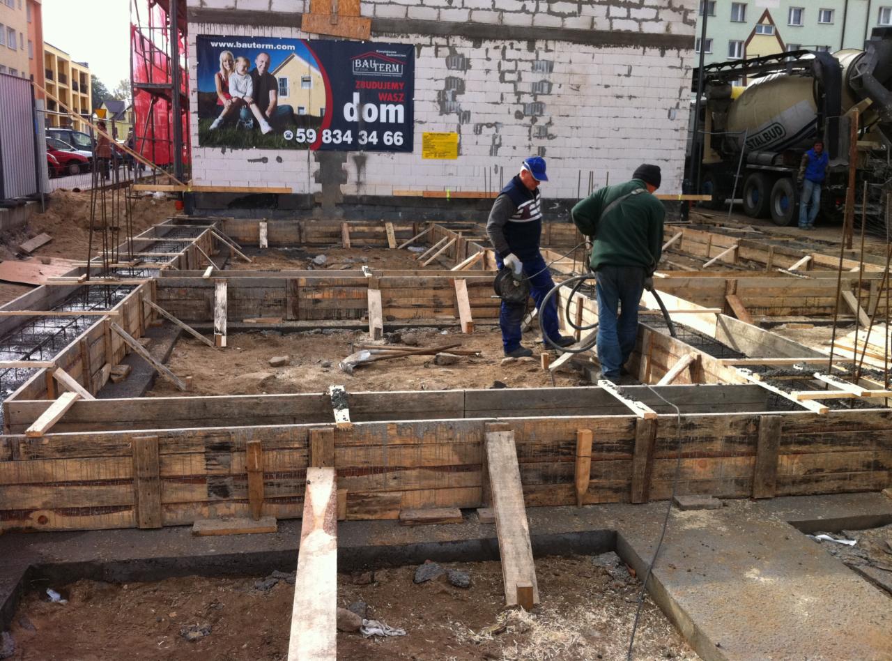 Wylewanie fundamentów z betonomieszarki w Człuchowie. Dwóch pracowników budowlanych w trakcie pracy. Szalunki drewniane i zbrojenie widoczne.