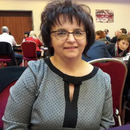 Portret uśmiechniętej kobiety w okularach, siedzącej przy stole podczas konferencji lub spotkania biznesowego, w tle widoczni inni uczestnicy.