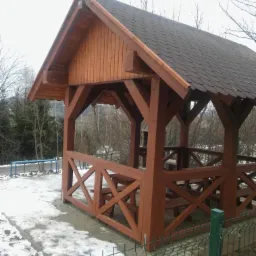 Drewniana altana z brązowym dachem krytym gontem bitumicznym, z widocznymi elementami konstrukcyjnymi i ławkami, otoczona śniegiem i siatką ogrodzeniową.