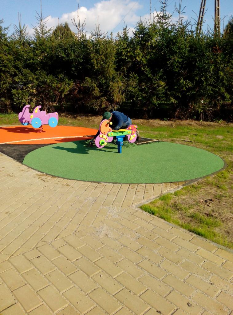 Plac zabaw z kolorowymi sprężynowcami w kształcie motocykli, jeden z nich użytkowany przez osobę w ciemnej kurtce, na tle zielonych krzewów i kostki brukowej.