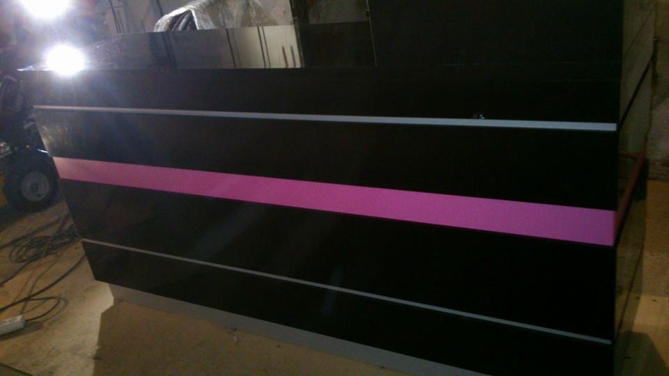 Częściowo widoczny, czarny mebel z poziomymi paskami w kolorze różowym i szarym, w tle widoczny sprzęt warsztatowy i nieporządek.