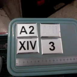 Cztery metalowe płytki z wygrawerowanymi oznaczeniami (A2, XII, XIV, 3) ułożone na plastikowej tacy, obok widoczna miarka i narzędzia. Precyzyjna obróbka.