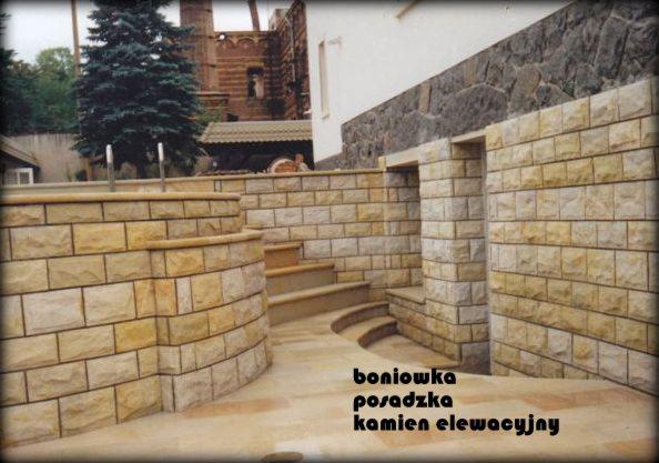 Elewacja budynku i schody wyłożone jasnym kamieniem elewacyjnym, z widoczną strukturą i nieregularnym ułożeniem, prowadzące do drzwi z kamienną ościeżnicą. W tle fragment budynku z ciemniejszego...