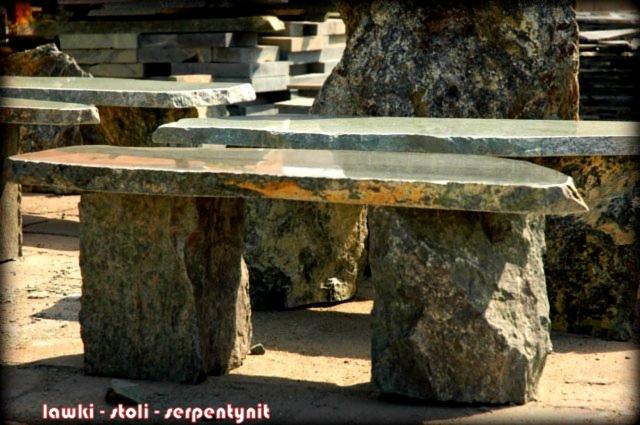 Surowy kamienny stół ogrodowy z blatem z serpentynitu i masywnymi nogami, prezentowany na tle stosu kamieni budowlanych.