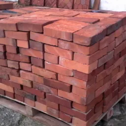 Paleta z cegłami w odcieniach czerwieni i pomarańczu, ułożonymi równo w stos, widoczne ślady użytkowania palety i zabrudzenia cegieł.