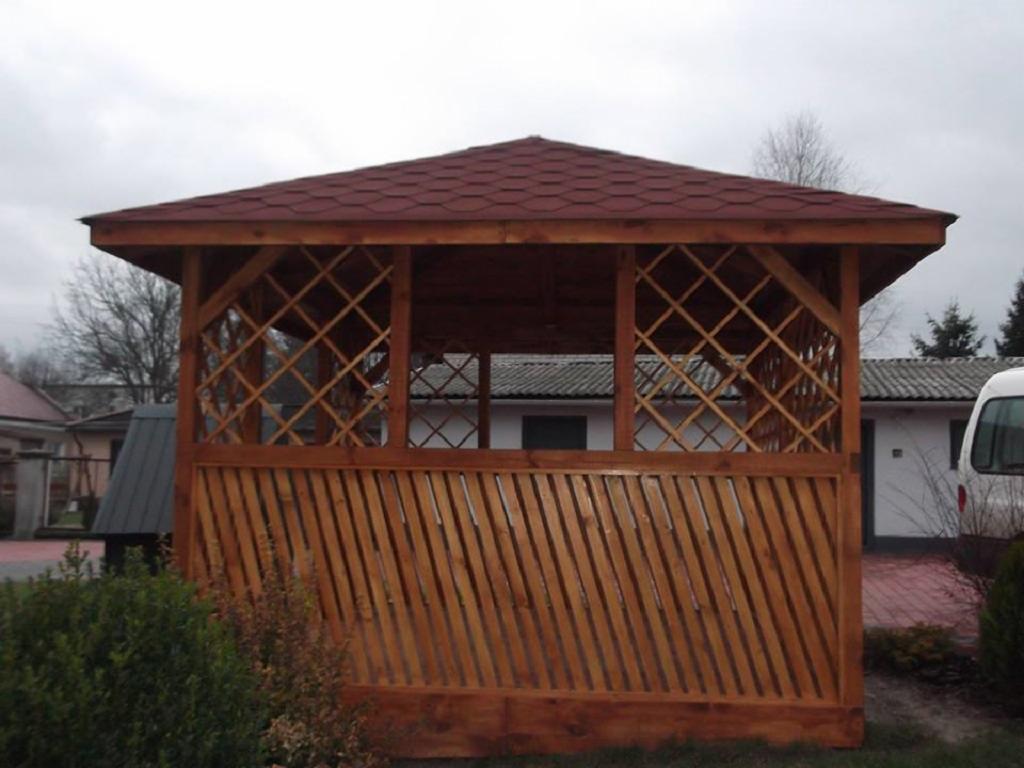 Drewniana altana z czerwonym dachem, kratowane ściany i ukośne deski, na tle zabudowań i pochmurnego nieba. Widoczny fragment krzewu.