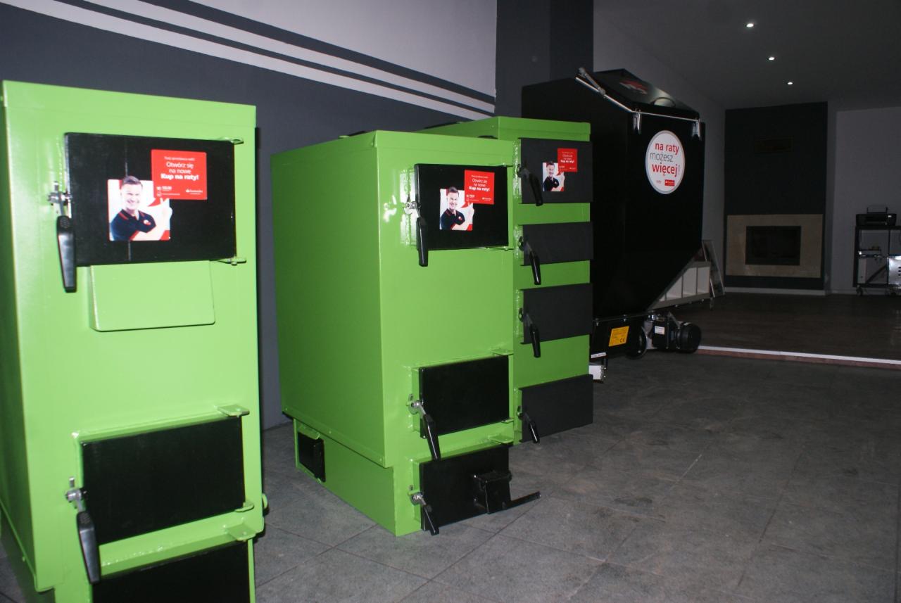 Zielone i czarne piece na paliwo stałe w salonie, z reklamami na drzwiczkach. Jeden z pieców ma napis 'na raty możesz więcej'.